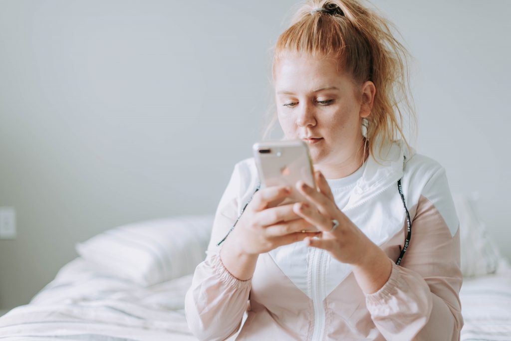Na zdjęciu widzimy młodą kobietę siedzącą na łóżku, która skupia się na swoim smartfonie. Kobieta ma długie, jasne włosy związane w kucyk i nosi jasną bluzę i białą koszulkę. W tle widoczne jest minimalistyczne wnętrze z jasnymi kolorami. Pościel na łóżku jest w odcieniach bieli. Kobieta wydaje się być zafascynowana tym, co widzi na ekranie telefonu, co sugeruje, że może przeglądać media społecznościowe, czytać wiadomości lub korzystać z aplikacji.