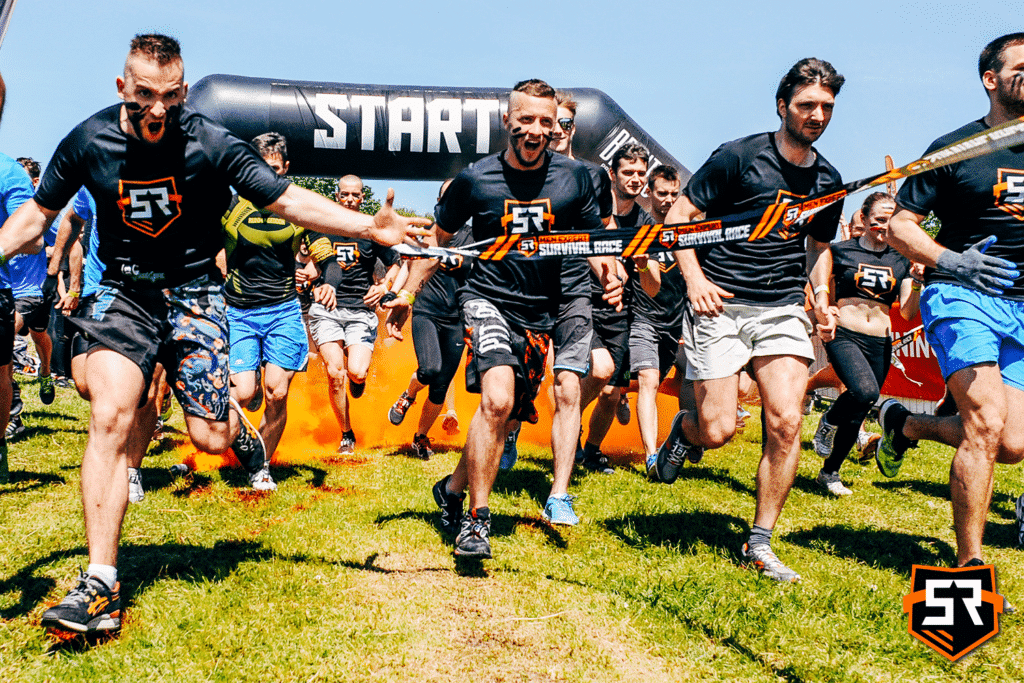 Na zdjęciu widzimy grupę uczestników biegu z przeszkodami „Survival Race.” Startują oni spod dużego, dmuchanego łuku oznaczonego słowem „START.” Uczestnicy ubrani są w sportowe stroje, a wielu z nich nosi koszulki z logo „Survival Race.” Wydarzenie odbywa się na otwartym terenie z trawiastą nawierzchnią, a nad nimi rozciąga się czyste niebo.