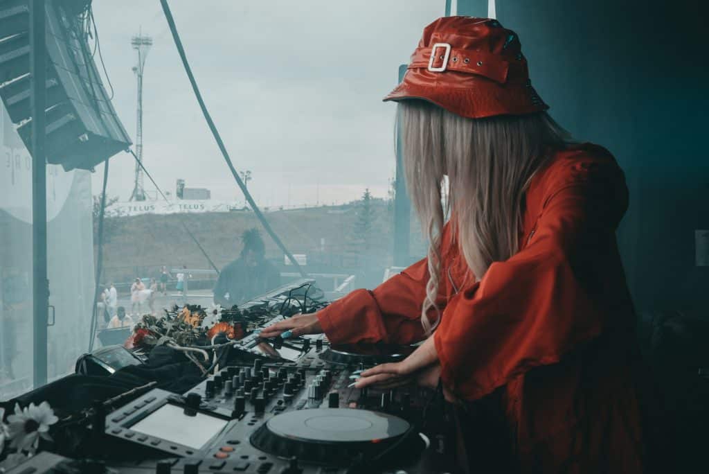 Na zdjęciu widzimy kobietę ubraną na czerwono, z czerwonym nakryciem głowy, obsługującą sprzęt DJ-ski. Ma długie blond włosy i dostraja ustawienia na konsoli DJ-a. Scena wydaje się być częścią wydarzenia plenerowego, ponieważ w tle widoczni są ludzie, a stanowisko DJ-skie znajduje się przy oknie lub otwartej przestrzeni. Sprzęt obejmuje różne pokrętła, przyciski oraz gramofon.