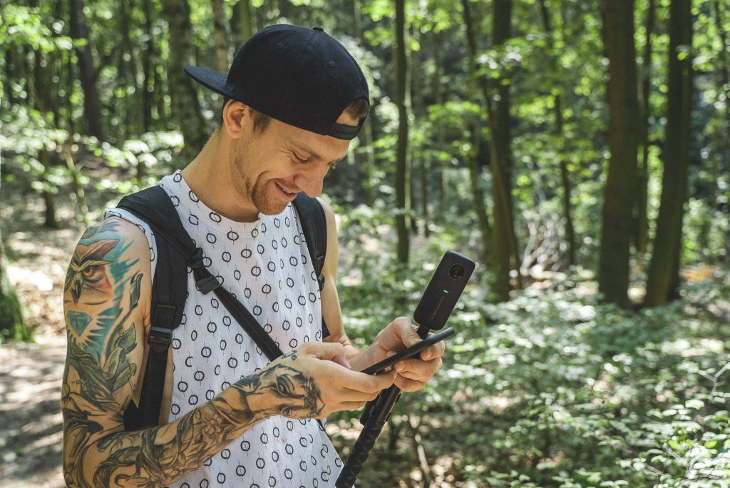 Zdjęcie przedstawia mężczyznę stojącego w leśnym otoczeniu. Ma na sobie białą koszulkę bez rękawów z okrągłym wzorem, czarną czapkę oraz czarny plecak. Na obu ramionach widoczne są tatuaże. Mężczyzna trzyma w jednej ręce smartfona, a w drugiej kijek do selfie. W tle widać drzewa i roślinność, co sugeruje, że może być na wędrówce lub przygodzie na świeżym powietrzu.