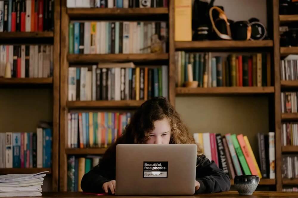 Zdjęcie przedstawia osobę o kręconych włosach siedzącą przy drewnianym stole, skupioną na pracy przy laptopie ozdobionym naklejką „Beautiful, free photos. Unsplash.” W tle rozciąga się duży regał pełen książek i dekoracyjnych przedmiotów, takich jak aparat fotograficzny czy imbryk, co nadaje wnętrzu osobisty, twórczy charakter. Kompozycja emanuje spokojem i koncentracją, typową dla domowego gabinetu lub biblioteki.
