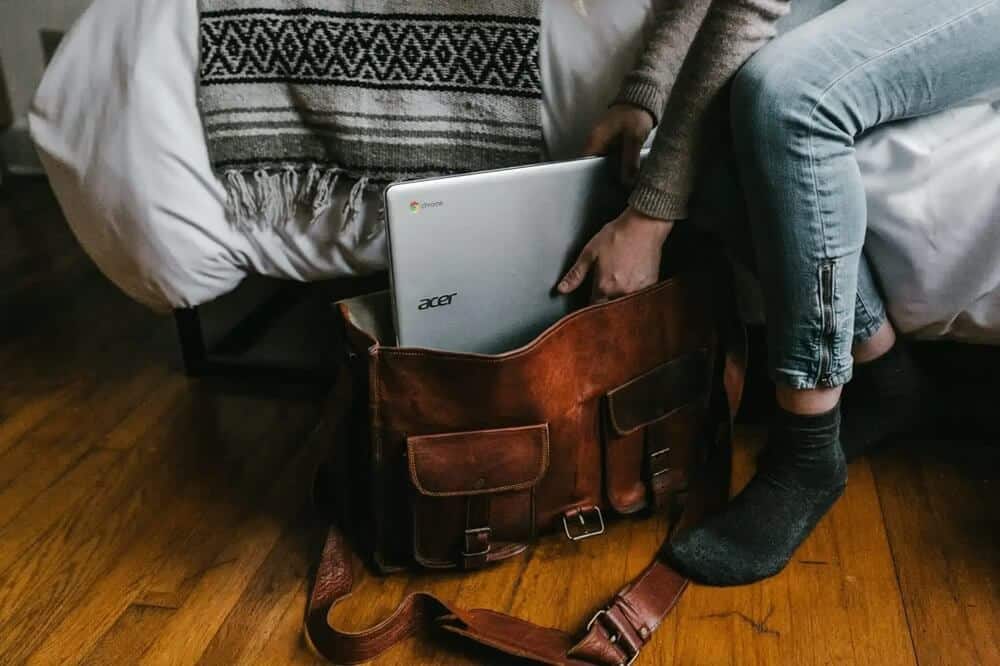 Zdjęcie przedstawia osobę siedzącą na brzegu łóżka, wkładającą srebrnego laptopa Acer Chromebook do brązowej skórzanej torby. Ubrana w brązowy sweter i jasnoniebieskie dżinsy, z widoczną stopą w ciemnej skarpetce, wygląda na gotową do wyjścia. Łóżko pokryte jest białą kołdrą, a na jego brzegu leży pleciony koc w czarno-białym wzorze, co dodaje wnętrzu przytulności. Drewniana podłoga i miękkie światło tworzą atmosferę domowego spokoju.